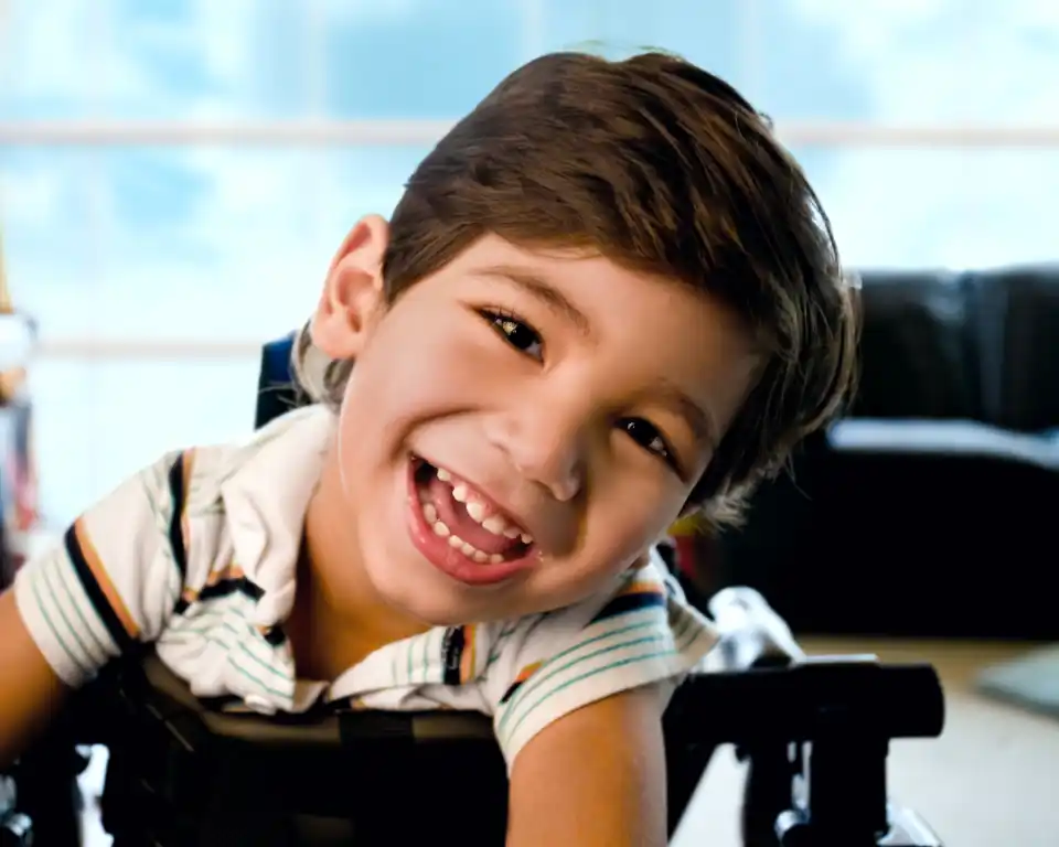 Smiling boy in adaptive chair, white striped polo, bright room with large windows — Prestige Health Partners pediatric care